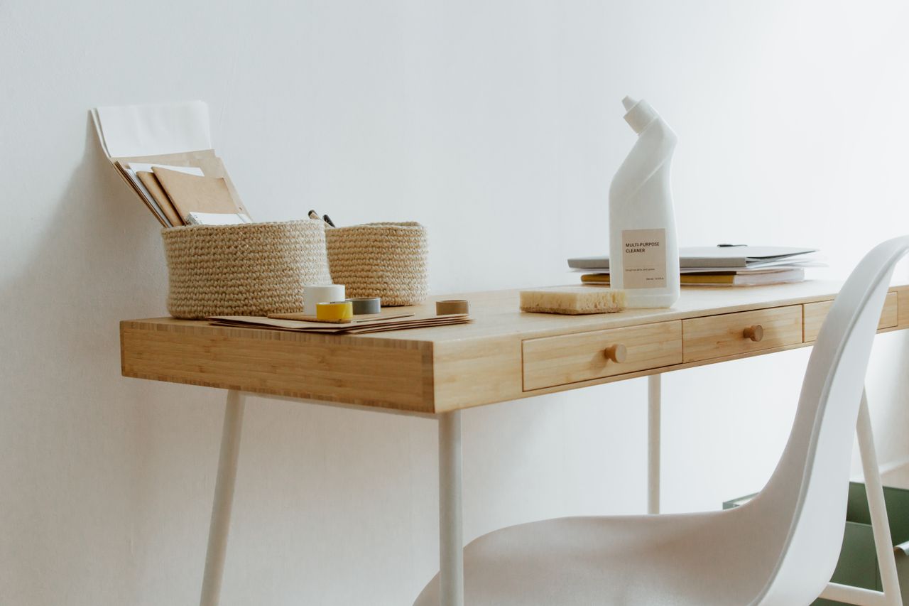 gallery-03 A clean and organized home office desk with cleaning products, papers, and storage baskets.